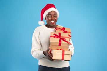 A cheerful woman wearing a Santa hat smiles while carrying several wrapped gifts with red ribbons. The background is a solid light blue, creating a festive atmosphere.