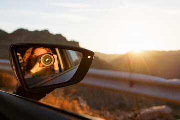 Reflection of a person taking a photo in a car side mirror during sunset.