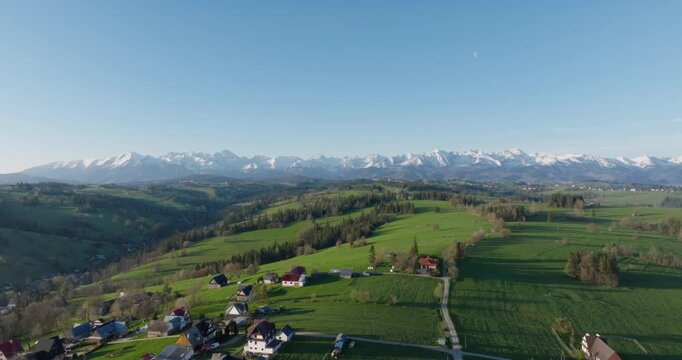Aerial view of a traditional mountain village in Podhale, surrounded by green hills and forests with snow-capped Tatra peaks. Highlights rural housing in a unique alpine climate.