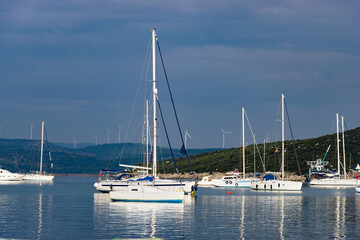 Obraz premium A tranquil marina filled with sailboats and yachts anchored on calm blue waters, with rolling green hills and distant wind turbines under a soft cloudy sky creating a peaceful coastal atmosphere.