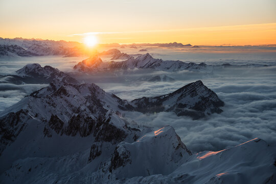 Aerial view of snow-dusted peaks pierce through a sea of clouds, illuminated by the radiant glow of the rising sun, Wildhaus-Alt Saint Johann, St. Gallen, Switzerland.