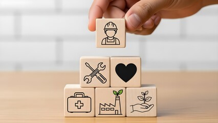 A hand placing a wooden block with a worker icon on top of a stack of blocks with various healthcare and business icons on a wooden table with a white brick wall background.