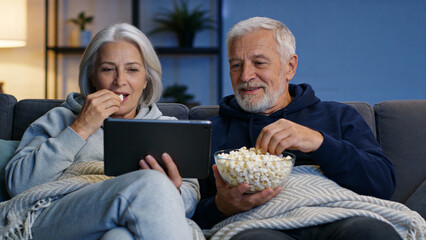 A senior caucasian woman and man eating popcorn while watching a movie on a digital tablet at home. Leisure and retired couple lifestyle.
