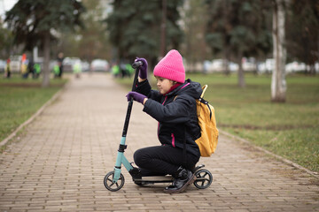 Obraz premium Cute little girl n a pink beanie rides an scooter along a tree-lined park path, wearing gloves and a backpack. Сhild is trying to regulate.