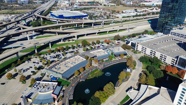 Interchange design facilitates seamless traffic movement across multiple levels, visible lane divisions, directional ramps, surrounding pondside commercial zones, wooden boardwalk, Plano, Texas