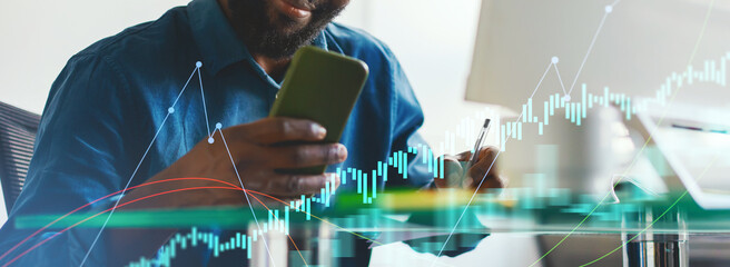 A man in a blue shirt analyzes market data on his smartphone while taking notes. He appears focused and engaged in his work at a modern office desk with digital graphics in view.
