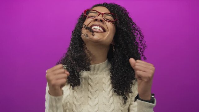 Woman wearing red glasses smiles and clenches fist in studio against purple wall; joy triumph success celebration victory happiness excitement elation euphoria bliss.