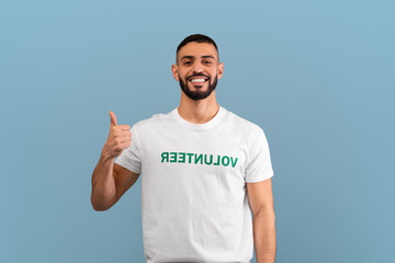 A young man wearing a white t-shirt with the word VOLUNTEER in green is smiling and giving a thumbs...