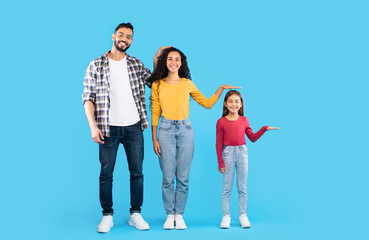 Fototapeta premium A joyful family poses against a bright blue backdrop, with a dad, mom, and young girl playfully comparing their heights. Each member shows a cheerful expression.
