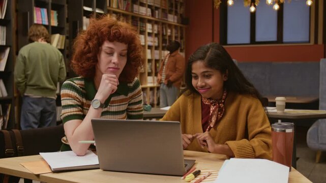 Young South Asian female student studying at library table with groupmate looking at laptop screen while browsing information and collaborating on academic research