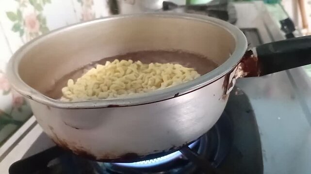 A block of instant noodles being dropped and slowly submerged into a pot of boiling water on a hot gas stove, showing the first steps of preparation for a quick meal.