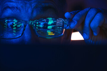 Man holding glasses while observing financial data reflecting on a monitor in dark room