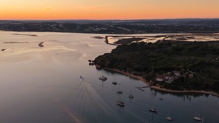 A tranquil sunset scene with boats anchored near a small sandy beach, a motorboat leaving ripples behind, and coastal wetlands and distant hills glowing under the orange sky.