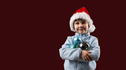 A little girl in a Santa hat, clutching Christmas tree baubles. A sign of Christmas and New Year's Eve. Copy space.