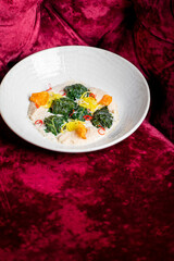 A gourmet dish featuring vibrant green leafy vegetables, a creamy white sauce, and colorful garnishes in a white bowl on a rich red velvet background.