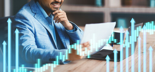 Indian businessman is deeply engaged in reviewing papers while seated at a desk with a laptop.