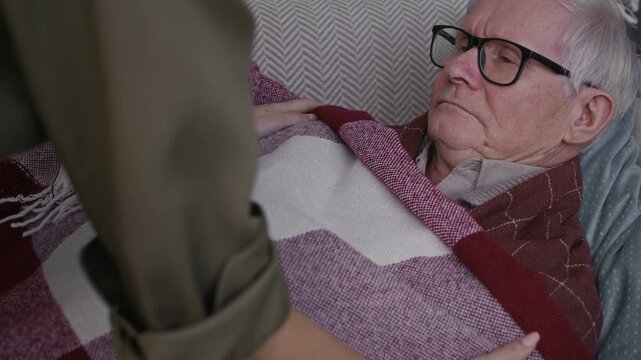 Medium shot of sick elderly man shivering on couch with fever getting body wrapped with blanket by female caregiver at home