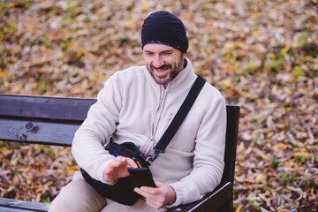 A man smiling while using his smartphone on a park bench in autumn. Relaxed outdoor activity, connectivity, and leisure in fall. © dizfoto1973