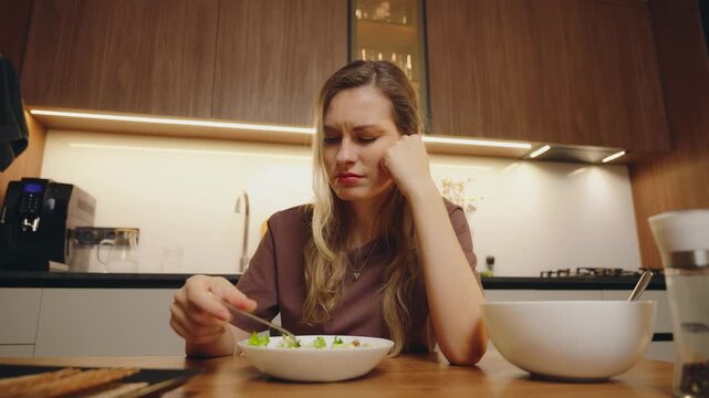 unhappy woman sitting at kitchen table preparing healthy vegetable salad bowl for diet dinner. Gigl mixing salad with fork on plate, bad appetite, sad expression refuses to eat greens healthy eating.