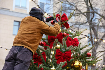 Man decorates the Christmas tree with red toy balls and bows. New Year decorations on winter street