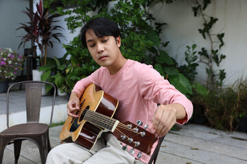Asian young man tuning acoustic guitar in outdoor garden seating area, relaxed musician adjusting string tension while seated