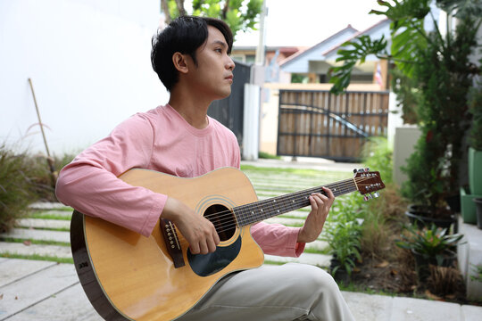 Asian Young man playing acoustic guitar outdoors in backyard patio, casual musician wearing pink long sleeve shirt, relaxed expression, natural light, home garden scene - Powered by Adobe