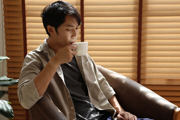 Asian young man sipping hot coffee in sunlight by wooden blinds, relaxed morning mood, casual shirt and armchair