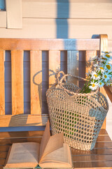 Summer season still life; open book, wicker bag with bouquet of white daisy flowers on a wooden bench by sunny summer day