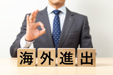 「海外進出」と書かれたブロックとOKポーズをするビジネスマン