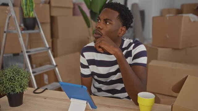 Young man holds tablet with finger on chin amid scattered cardboard boxes and potted plant on table in a building; contemplation.