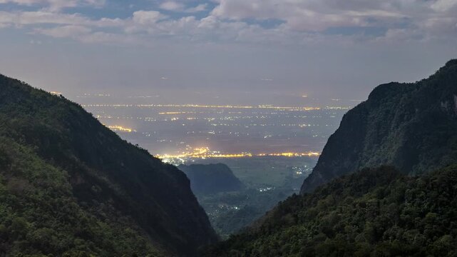 City lights illuminate the Pha Hee valley and Chiang Rai urban area nestled between dark, forested mountains in Asia. Stunning night scene captured during a cold twilight for travel and geography phot