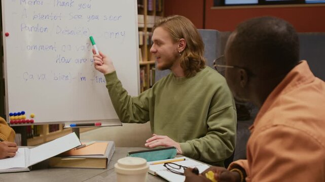 Friendly male language tutor presenting basic French phrases pointing at whiteboard while young adult Black man writing down notes in group elementary level class in library