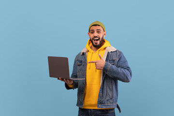 A cheerful young man is wearing a yellow hoodie and a denim jacket. He is holding a laptop and pointing at it with enthusiasm against a blue background.