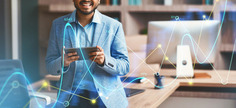 A confident Indian businessman with eyeglasses is smiling while browsing on a digital tablet in a modern office. He stands in front of a stylish workspace filled with business elements.