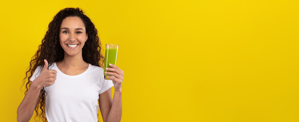 A young woman with curly hair is giving a thumbs up while holding a glass of green juice. She stands in front of a bright yellow background, radiating happiness and positivity.