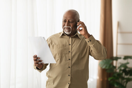 A senior man is standing in a bright room, holding a sheet of paper and talking on his phone. He appears focused and concerned, with sunlight filtering through the curtains. - Powered by Adobe