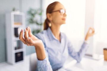 A relaxed woman sits cross-legged at her desk in a modern office, practicing mindfulness. Soft sunlight filters through the windows, creating a serene working environment.