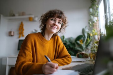 Young woman wearing glasses orange sweater smiling at camera writing in notebook at home workspace with laptop and plants