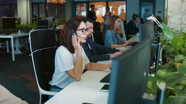 Female wearing headset sitting in busy office workspace. Listening to client while typing on keyboard. Colleagues focused on computers nearby. Support team handling communication in dim office.
