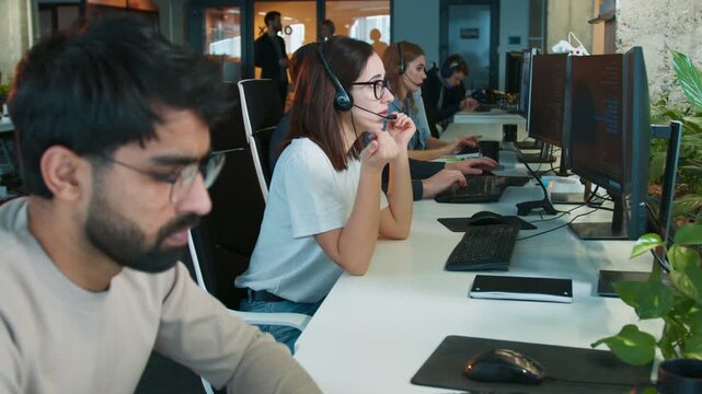 Female wearing headset sitting in busy office workspace. New worker getting nervous while communicating with client. Colleagues focused on computers nearby. Support team handling calls.