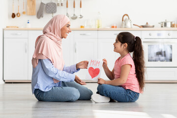 A young girl and her mother happily sit on the floor in a warm kitchen, crafting a colorful card that reads Mom with a red heart. Their smiles showcase a joyful moment of creativity.