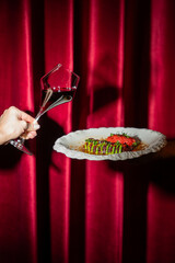 A hand holds a unique glass of red wine with a creative angle next to an elegant plate of gourmet...