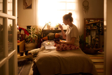 Caucasian young adult woman sitting cross legged on bed eating breakfast while working on laptop in cozy bedroom, with plants and personal items visible in background