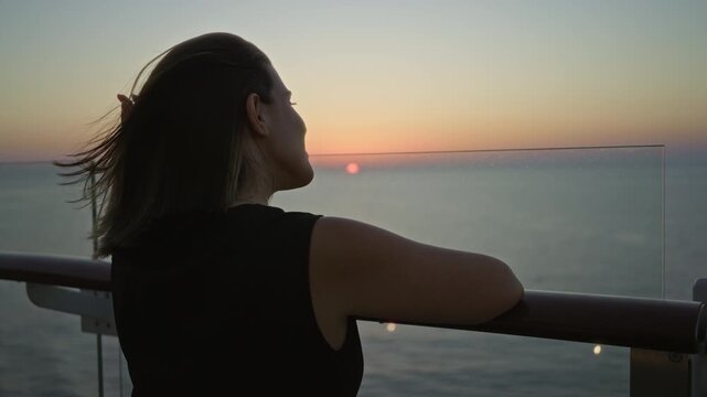 Woman leans on cruise ship rail, looking toward the horizon, showing a bare shoulder and resting hand to neck on deck at sunset; serenity.
