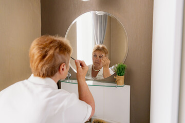 An elderly woman paints her eyebrows while looking in the mirror.