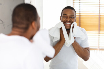 A man stands in front of a bathroom mirror, happily drying his face with two white towels. Sunlight streams through the window, creating a warm atmosphere.