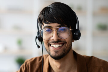A young man with glasses and a headset smiles while engaged in a conversation at his home office....
