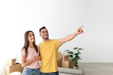 A young couple stands together in their new home filled with cardboard boxes. They are smiling,...