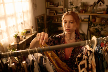 Young adult Caucasian woman selecting clothing from rack in sunlit room, focused expression on...