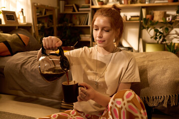 Caucasian young adult woman sitting on floor pouring coffee from glass pot into mug in cozy living room, relaxed expression, long hair tied back, surrounded by books and plants
